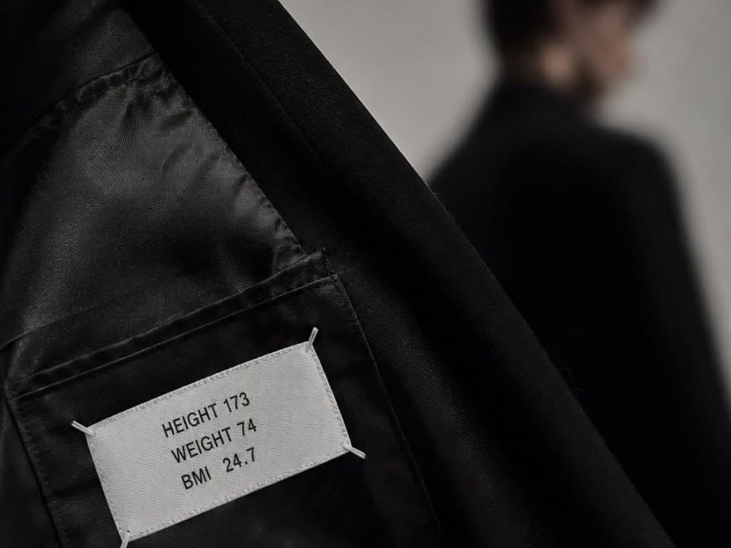 A fabric label stitched inside a black jacket reading HEIGHT 173, WEIGHT 74, BMI 24.7, with a softly blurred figure standing in the background