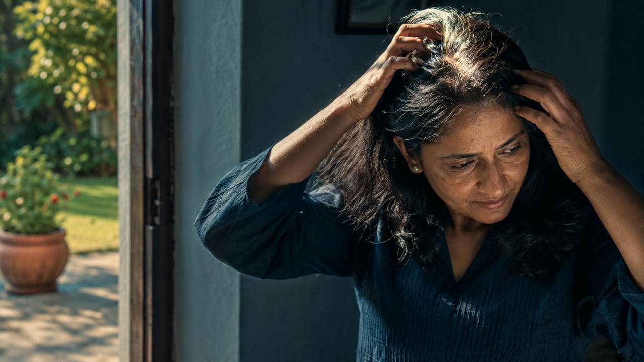 An Indian woman in her forties parting her hair with both hands to check her scalp in natural morning light, a garden visible through the doorway behind her