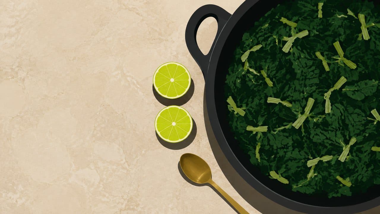 A flat-lay on beige marble: a black cast-iron pan of cooked dark leafy greens, two lime halves cut side up beside it, and a long gold spoon resting alongside the pan.