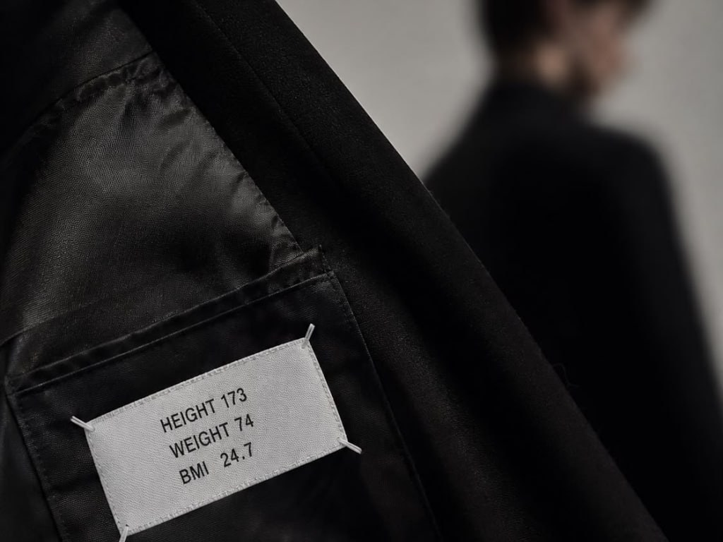 A fabric label stitched inside a black jacket reading HEIGHT 173, WEIGHT 74, BMI 24.7, with a softly blurred figure standing in the background
