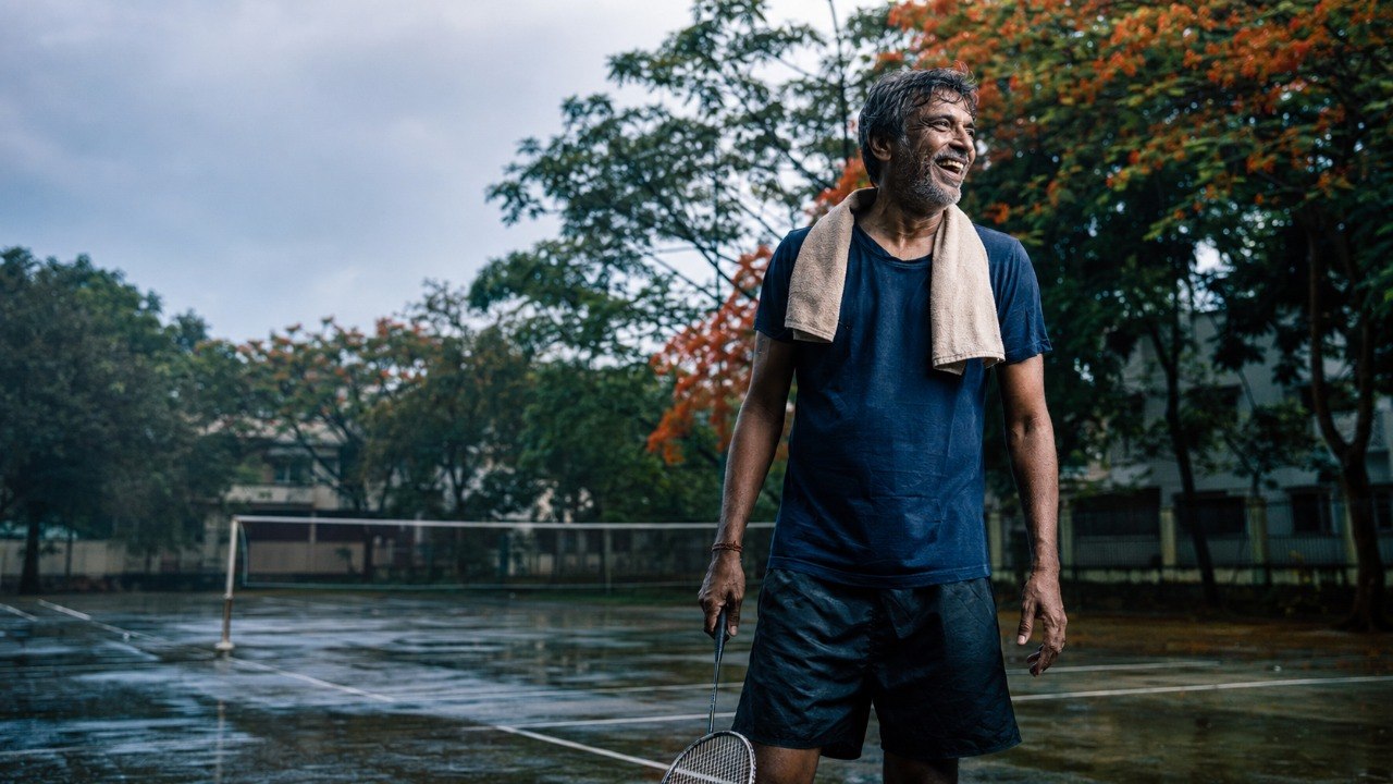 A middle-aged Indian man laughing on a wet badminton court after rain, a towel around his neck and a racquet in hand