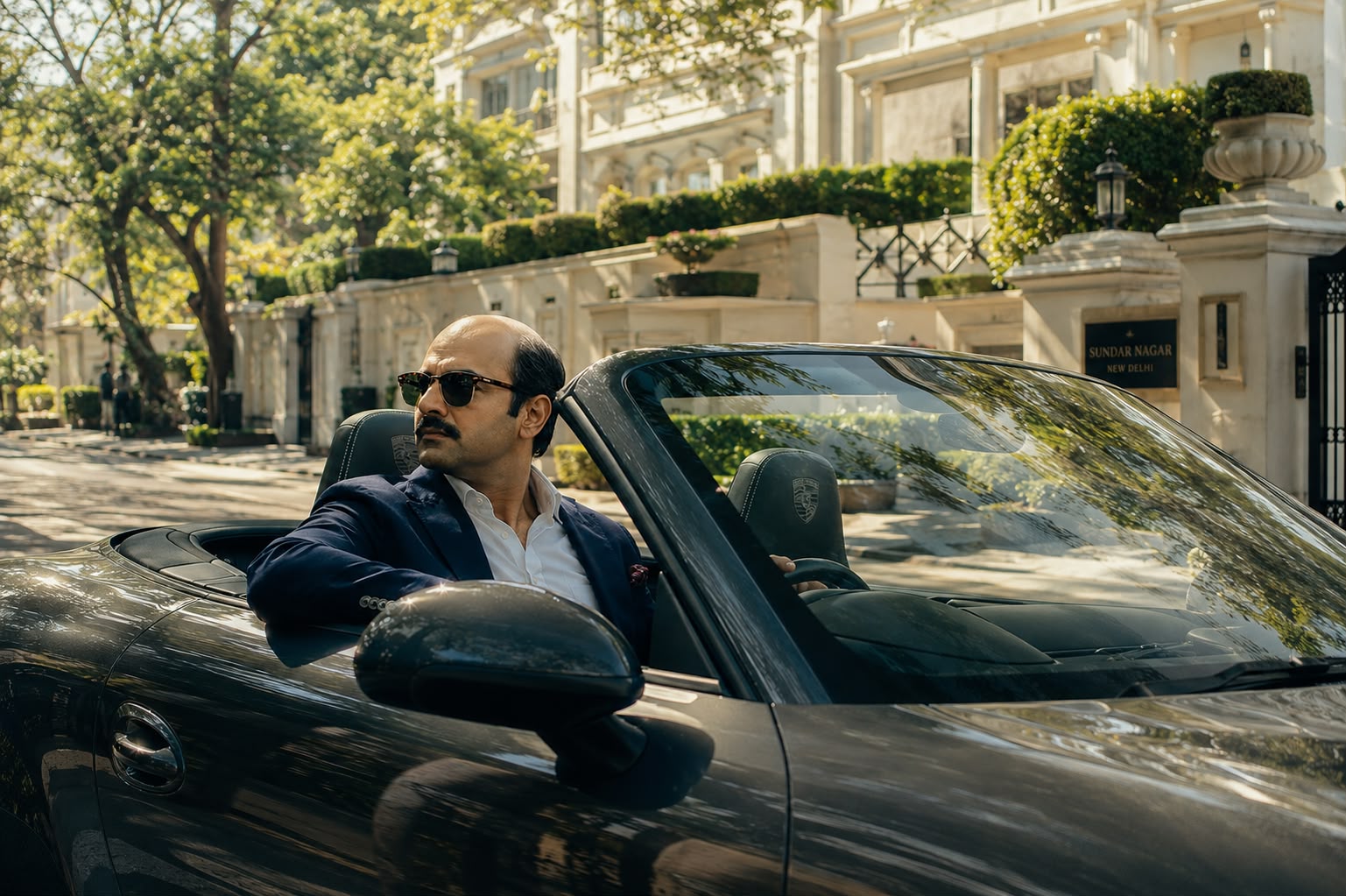 A balding Indian man in sunglasses and a navy blazer sitting in an open-top car on a tree-lined street in Sundar Nagar, New Delhi
