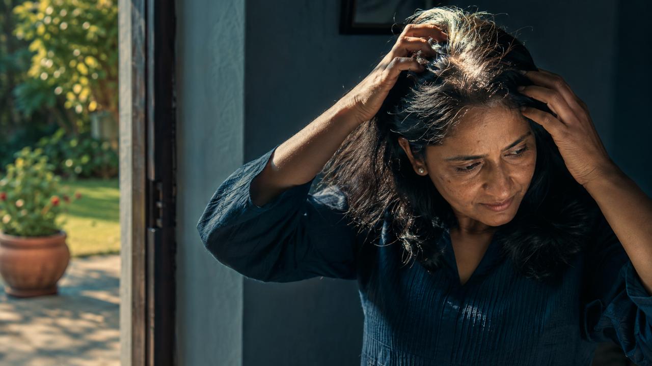 An Indian woman in her forties parting her hair with both hands to check her scalp in natural morning light, a garden visible through the doorway behind her