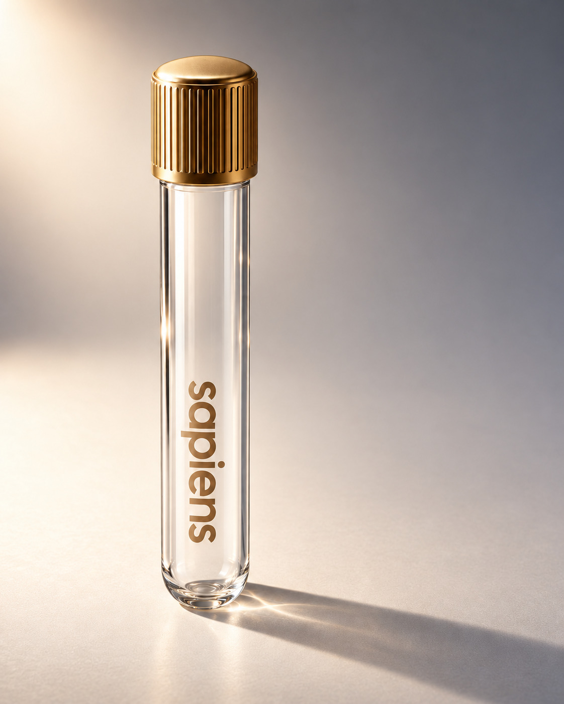 A Sapiens-branded glass vacutainer with a fluted gold cap standing upright on a pale cream surface in soft directional daylight, its shadow falling to the right.